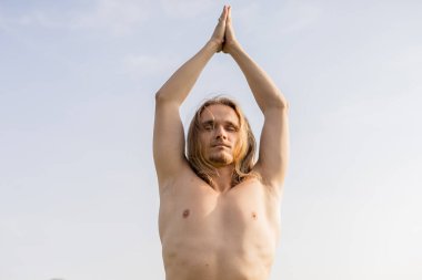 Uzun saçlı, gözleri kapalı, mavi gökyüzüne karşı dua eden ellerle meditasyon yapan üstsüz bir adamın alçak açılı görüntüsü.