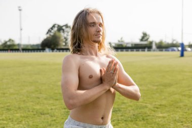 Üstsüz, uzun saçlı yoga adamı çimenli stadyumda dua eden elleriyle meditasyon yapıyor.