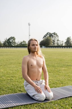Açık hava stadyumunun yeşil sahasında, gök gürültüsü pozunda meditasyon yapan keten pantolonlu, pozitif tişörtsüz bir adam.