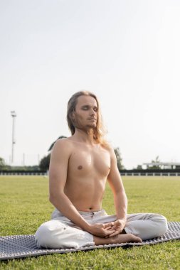 Üstsüz ve sportif gözleri kapalı bir adam yeşil çimlerde rahat yoga pozunda meditasyon yapıyor.
