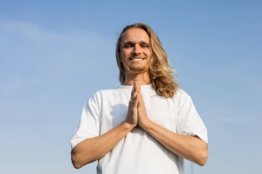 Düşük açılı uzun saçlı adamın mavi bulutsuz gökyüzüne karşı Anjali Mudra jesti ile meditasyon yapması.
