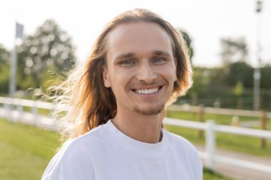 portrait of pleased long haired yoga man in white t-shirt smiling at camera outdoors clipart