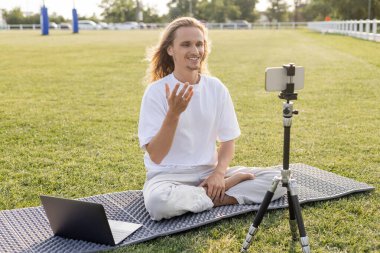 Mutlu yoga koçu rahat bir şekilde dizüstü bilgisayar ve cep telefonuyla dışarıda stadyumun yeşil çimlerinde poz veriyor.