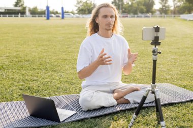 Yoga koçu akıllı telefon ve dizüstü bilgisayarla tripodun yanında çimenli stadyumda rahat poz verirken konuşuyor.