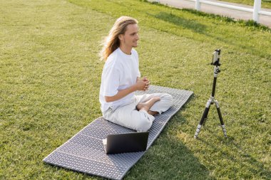 Gülümseyen yoga hocası tripodun yanında yeşil çimlerde akıllı telefon ve dizüstü bilgisayarla poz veriyor.