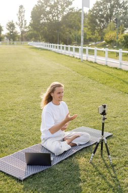 joyful yoga coach pointing with hand near smartphone and laptop on outdoor stadium clipart