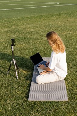 long haired yoga teacher using laptop while sitting near tripod with cellphone on grassy stadium clipart
