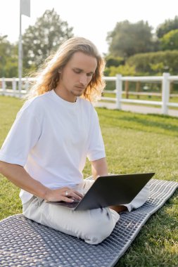 Genç yoga adamı çimenli stadyumda rahat poz veriyor ve online ders sırasında dizüstü bilgisayar kullanıyor.