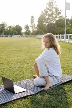 Uzun saçlı, adaçayı rengi poz veren bir adam online ders sırasında açık hava stadyumunun yeşil çimlerinde dizüstü bilgisayarda poz veriyor.