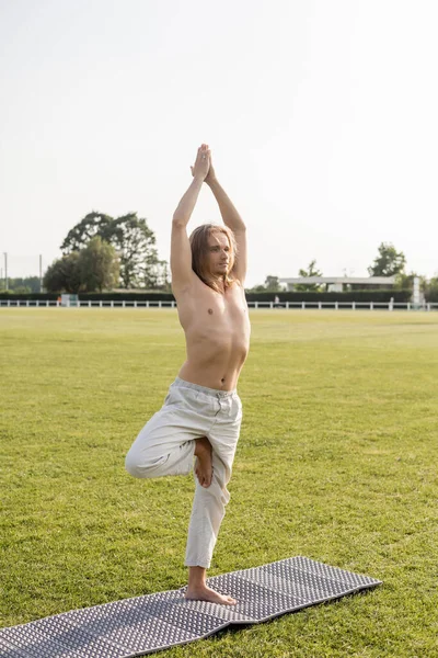 shirtless barefoot man in linen pants meditating in tree pose with raised hands on yoga mat outdoors