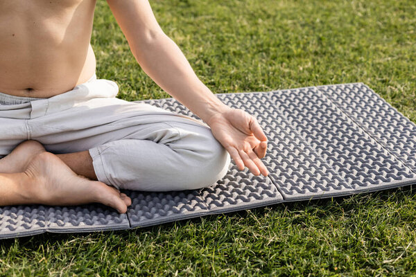 частичный взгляд человека без рубашки в льняных брюках медитируя в легкой позе с жестом мудра gyan на циновке yoga на зеленой траве