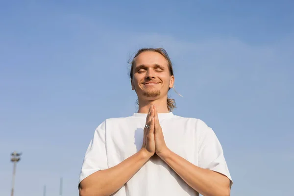 pleased man in white cotton t-shirt meditating with closed eyes and anjali mudra gesture against clear sky