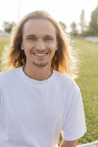 портрет счастливого длинноволосого мужчины в белой хлопчатобумажной футболке, улыбающегося в камеру на улице