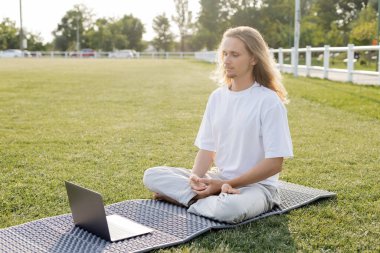 Genç adam yoga minderinde dizüstü bilgisayarın yanında meditasyon yapıyor. 
