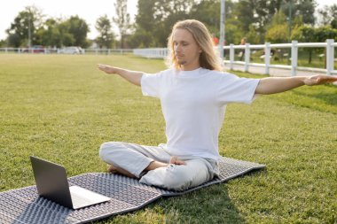 Genç adam açık havada dizüstü bilgisayarda yoga dersi sırasında ellerini uzatarak poz veriyor.