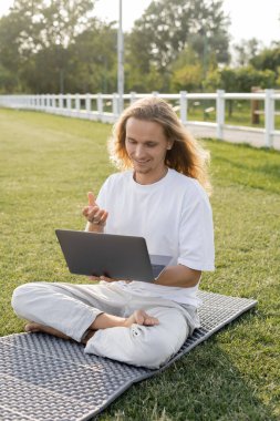 Yoga dersinde gülümseyen adam dışarıda stadyumda rahat poz verirken dizüstü bilgisayarda konuşuyor.