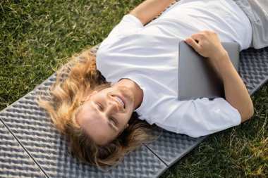 Yeşil çimlerin üzerinde laptopuyla yoga minderinde uzanan kaygısız, uzun saçlı adamın üst görüntüsü.