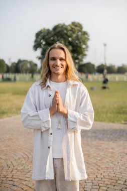 Uzun saçlı yoga adamı kameraya bakıyor ve dışarıda dururken Anjali Mudra hareketi gösteriyor.