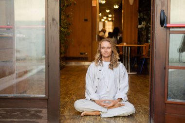 Beyaz keten elbiseli çıplak ayaklı bir adam kulübenin girişinde rahat bir şekilde meditasyon yapıyor.