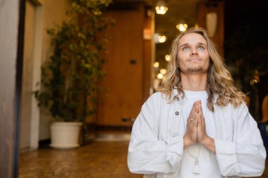 Beyaz gömlekli şık, uzun saçlı bir adam. Anjali Mudra jesti yapıyor ve evde meditasyon yaparken kafasını kaldırıyor.