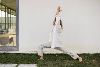 Beyaz pamuklu giysiler içinde çıplak ayaklı bir adamın savaşçı pozunda meditasyon yaparken modern kulübenin yakınında dua eden eller.