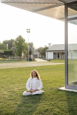 Nilüfer çiçekli beyaz elbiseli hipster adam kır evinin yakınındaki yeşil çimlerde meditasyon yaparken poz veriyor.