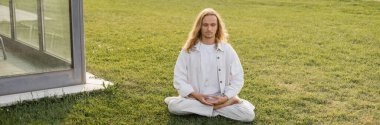 young man in white clothes meditating in lotus pose on green lawn near building, banner clipart