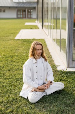 Uzun boylu, beyaz elbiseli bir adam. Evin yakınındaki çimlerde otururken lotus pozisyonunda yoga yapıyor.