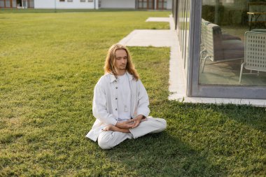 Nilüfer çiçeği pozunda meditasyon yapan şık, uzun saçlı bir adam binanın yanındaki çimenlikte otururken.