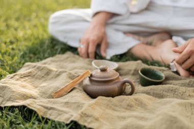 Yoga yapan adamın palo santo sopasıyla keten halının yanında oturması ve yeşil çimenlikte fincanlı kil çaydanlığı.