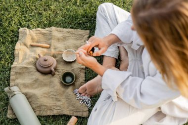 Yogacı adamın elinde kokulu sopayla kil çaydanlık ve çimenli çimlerde kâselerle keten halıların yanında.