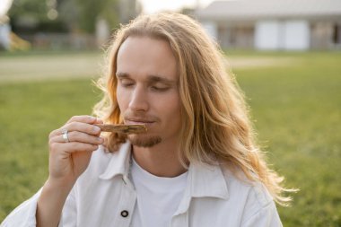 long haired yoga man with closed eyes enjoying flavor of palo santo stick outdoors clipart