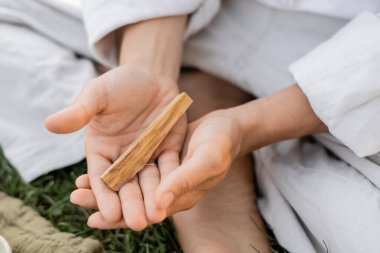 Beyaz keten elbiseli bir adam dışarıda otururken elinde aromatik palo santo tutarken kısmen görülüyor.