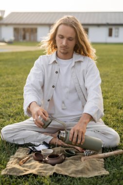 long haired man in linen clothes pouring hot water from thermos into ceramic teapot  clipart