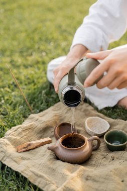 Yogacı adamın seramik çaydanlığa sıcak su dökerken keten halı ve palo santo sopasıyla çimenli çimlerin üzerinde oturduğu görüntüler.