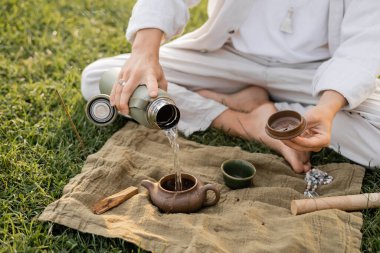 Yogacı adamın kil çaydanlığa sıcak su dökerken, keten halıların yanındaki çimlerde otururken, mala boncukları ve palo santo çubuğuyla kısmi görüntüsü.