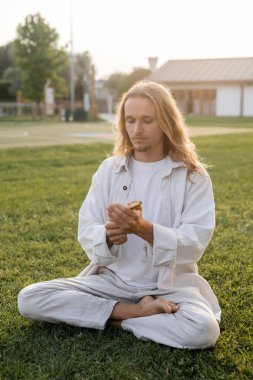 Beyaz elbiseli şık, uzun saçlı adam rahat bir şekilde meditasyon yapıyor ve yeşil çimlerde palo santo sopası tutuyor.