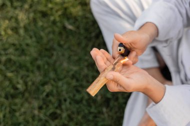 partial view of yoga man sitting on green lawn and lightening fragrant palo santo stick clipart