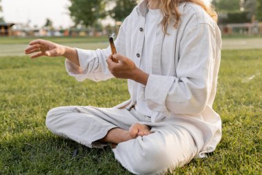partial view of man in white clothes holding smoldering palo santo stick and meditating in easy pose on green lawn clipart