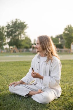 Beyaz pamuklu giysiler içinde kaygısız bir adam açık yeşil çimenlere yapışmış kokulu palo santo ile rahat bir şekilde meditasyon yapıyor.