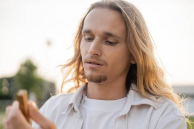 portrait of young man with long fair hair holding blurred palo santo stick outdoors clipart