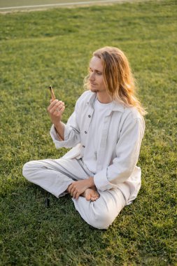 young yoga man in white clothes looking at fragrant palo santo stick while sitting in sukhasana pose on green grass clipart