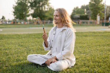 Mutlu, uzun saçlı, elinde aromatik palo santo tutan adam yeşil çimlerde rahat bir şekilde meditasyon yapıyor.