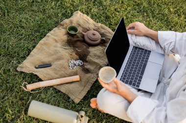 partial view of man with laptop holding clay cup near teapot and mala beads on linen rug on green lawn clipart