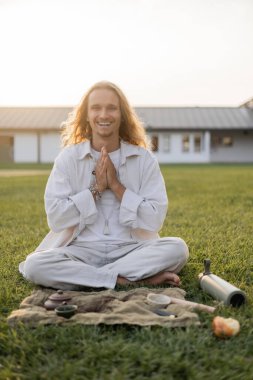 Keten elbiseli kaygısız bir adam kırsaldaki yeşil çimlerde çay seremonisi sırasında kil bardakların ve çaydanlıkların yanında meditasyon yapıyor.