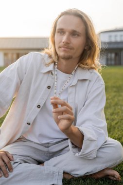 long haired man in linen clothes touching mala beads and looking away during meditation outdoors clipart