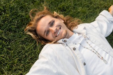 high angle view of cheerful young man in white linen shirt lying on green lawn and smiling at camera clipart