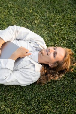 top view of overjoyed man with long fair hair holding laptop while lying on grassy field