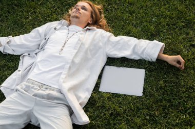 Yeşil tarlada dizüstü bilgisayarın yanında yatan beyaz elbiseli genç yoga adamı yüksek açılı görüyor. 