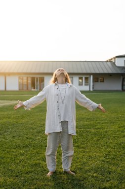 Uzun boylu, keten elbiseli bir adam uzanmış elleriyle meditasyon yapıyor ve kır evinin yakınındaki yeşil çimlerde başını kaldırıyor.